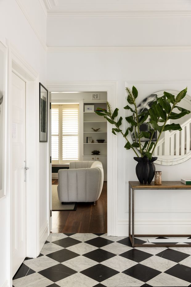 Elegant residential hallway or entry foyer with a dramatic black-and-white checkerboard tile floor, white paneled walls, ornate crown moulding, and a narrow timber-topped console table. A large leafy plant and rounded wall mirror sit above the console. Through the open doorway is a refined living room with a pale upholstered armchair, dark timber flooring, built-in shelving, and plantation shutters. The interior is bright, orderly, and classically detailed.