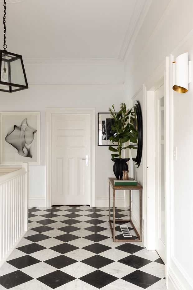 A bright, refined residential hallway or landing with white paneled walls, decorative ceiling cornices, and a black-and-white checkerboard floor. A console table with a large potted plant, round mirror, and framed artwork lines the wall, while a black geometric pendant and contemporary wall sconces provide lighting. A staircase balustrade is visible at left.