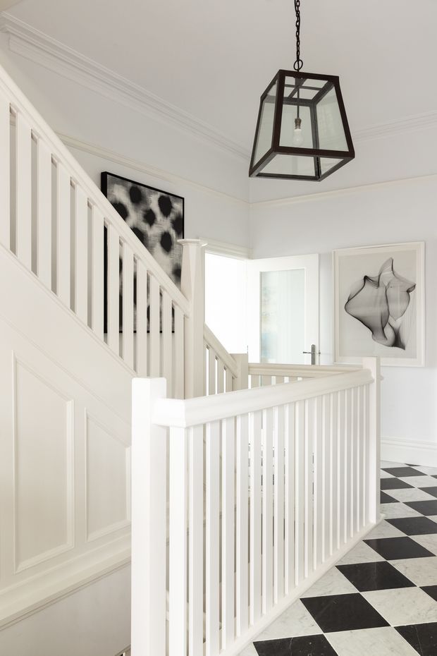 Bright, refined residential stair landing with white painted timber panelled walls, traditional moulded cornices and trim, and a white balustrade. The floor features a bold black-and-white checkerboard pattern, complemented by a dark metal and glass lantern pendant. Framed monochrome artwork and a glazed doorway add decorative detail.