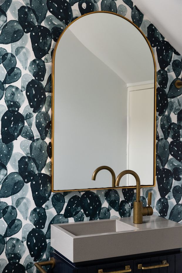 Close-up of a stylish bathroom vanity featuring a large arched mirror with a slim brass frame, a wall-mounted rectangular basin, and brass tapware. The walls are covered in bold dark green and blue botanical-patterned wallpaper, with dark cabinetry and brass hardware below the basin.