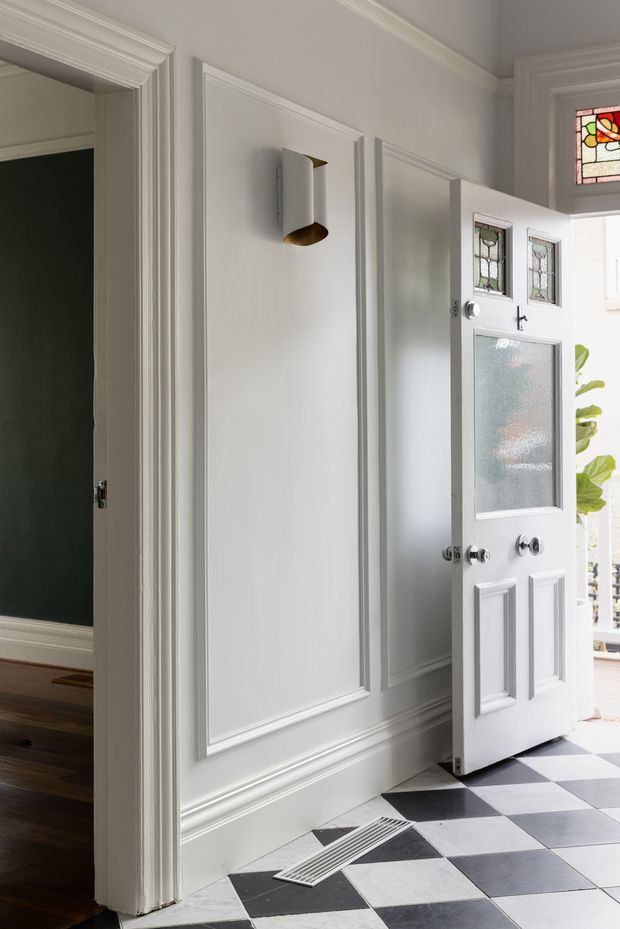 A bright, refined residential entry hall with white decorative wall panelling, substantial moulded door and skirting trims, and a partially open front door featuring leaded-glass panels and frosted glazing. The floor is finished in a bold black-and-white checkerboard pattern, with a sculptural wall sconce and a glimpse of an adjacent dark-painted room. Traditional detailing is balanced by clean contemporary lighting and hardware.