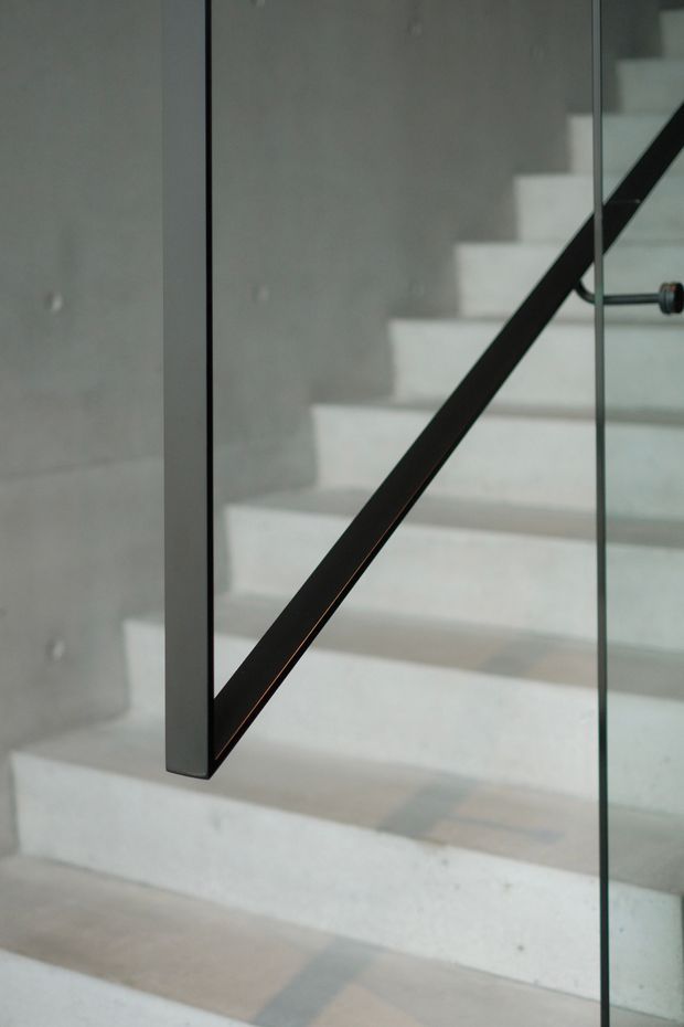 Close-up architectural detail of a minimalist stairway with pale concrete treads and a smooth concrete wall. A clear glass balustrade is supported by a slim black metal handrail and vertical frame, creating a stark, contemporary geometric composition.