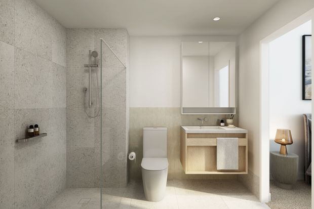 A bright, contemporary bathroom featuring a walk-in glass shower with large pale stone-look wall tiles, a wall-mounted toilet, and a floating timber vanity with an integrated basin beneath a mirrored cabinet. A doorway opens to an adjacent furnished room, and natural light enters from the right.