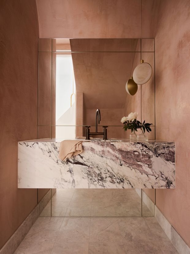A compact, highly refined bathroom featuring a floating double vanity clad in dramatic veined marble, integrated bronze-toned tapware, a large wall-to-wall mirror, and warm pink-toned plaster walls. Matching stone tiles extend across the floor and lower wall edges, while sculptural pendant lights and a small floral arrangement add a luxurious decorative finish.