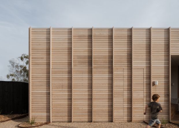 Exterior view of a contemporary single-storey building facade clad in vertically divided panels of horizontal light timber boards. The repetitive timber screen creates a strong linear rhythm, with a recessed entry at the right and a dark boundary fence and sparse vegetation surrounding the building.