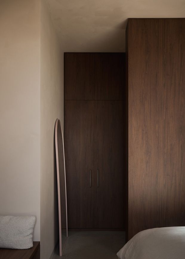 A moody, minimalist bedroom corner with dark timber built-in wardrobes, a narrow full-length mirror, warm off-white plaster walls, and part of a bed visible in the foreground. The concealed wardrobe joinery and restrained palette create a calm, refined atmosphere.