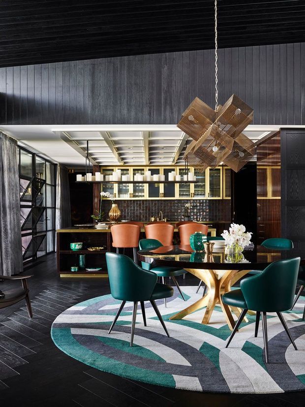 A dramatic, richly layered open-plan dining and home bar interior. The room features dark timber wall panelling and flooring, a coffered white ceiling, black-framed glazing, a geometric sculptural pendant light, a built-in bar with illuminated shelving, teal upholstered dining chairs, rust-orange bar stools, a polished gold-toned dining table base, and a large abstract geometric rug in teal, grey, white, and black.
