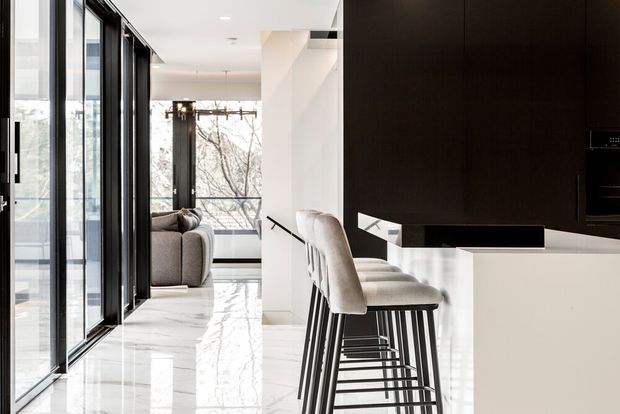 Bright contemporary open-plan kitchen and living area viewed along a polished light floor. The kitchen features a white island with a dark stone-looking benchtop, tall dark cabinetry, integrated appliances, and a row of upholstered bar stools. Black-framed floor-to-ceiling glazing lines the left side, while a living area and staircase are visible beyond. The space has strong contrast between white surfaces and dark joinery and frames, with abundant natural light.