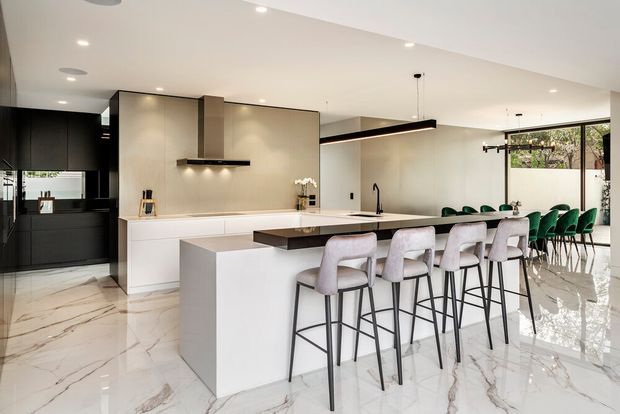 A spacious, contemporary open-plan kitchen and dining area with a large white island, integrated white cabinetry, dark accent cabinetry, and a sleek black linear pendant light. The kitchen features a pale stone-look backsplash, dark tapware, and a long island with several upholstered bar stools. A dining table with green upholstered chairs sits behind the island. Large windows bring in natural light, while the polished white veined floor creates a luxurious, reflective finish.