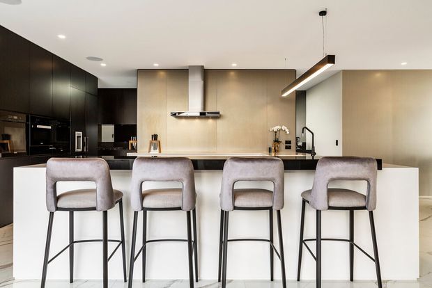 Contemporary open-plan kitchen viewed from the breakfast bar, with four upholstered bar stools, a dark full-height kitchen wall containing integrated appliances, a central island with pale stone surfaces, warm beige cabinetry or wall panels, black fixtures, and a linear pendant light. The room has a clean, refined, high-end appearance with recessed ceiling lighting and light flooring.