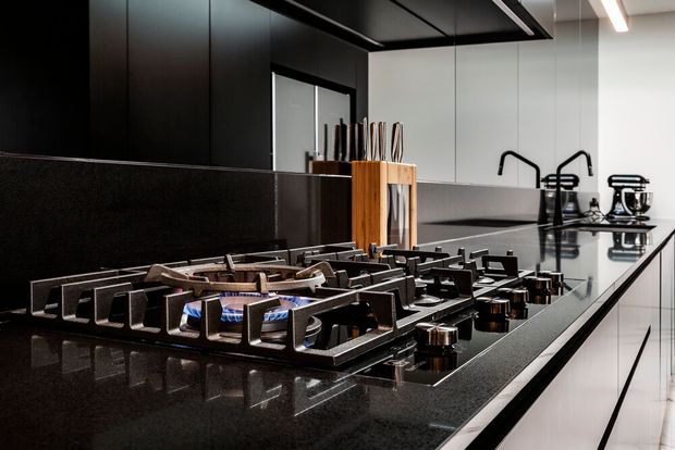Close-up view of a sleek contemporary kitchen centred on a large black gas cooktop set into a polished dark stone island benchtop. Minimal white and black cabinetry, a reflective black splashback, modern black tapware, integrated appliances, a timber knife block and linear ceiling lighting create a high-contrast, refined kitchen interior.