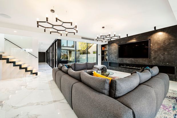 A spacious contemporary living room with a large grey sectional sofa arranged around a low coffee table, facing a wall-mounted television and linear fireplace set into a dramatic dark textured feature wall. Glossy white marble-look flooring, extensive glazing, black-framed internal partitions, open shelving, a sculptural geometric chandelier, and a floating staircase contribute to the luxurious modern interior.