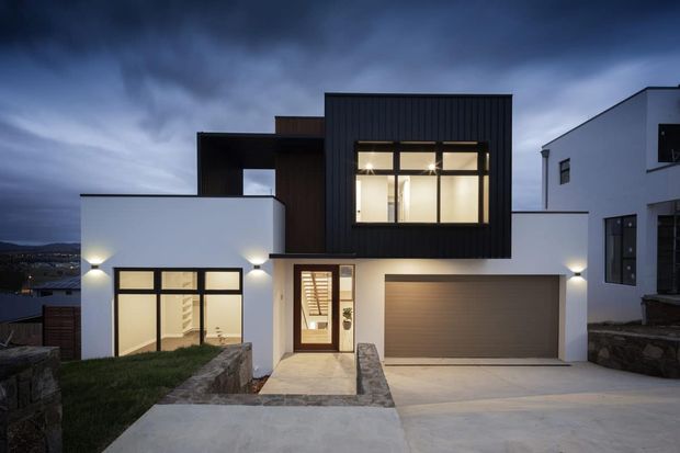 Contemporary two-storey residential home photographed at dusk, featuring a crisp white rendered facade, dark vertically ribbed upper volume, large black-framed windows, recessed entry, attached double garage, and layered flat-roofed forms. A sloping driveway and low stone retaining walls frame the front elevation, with warm interior and exterior lighting creating contrast against the dark sky.