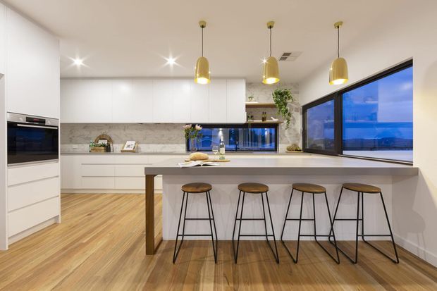Contemporary kitchen with white full-height cabinetry, a marble-look splashback, timber flooring, and a long island bench with seating for four. Three pendant lights hang above the island, while large black-framed corner glazing brings in blue evening light. Integrated appliances and a small display shelf complete the bright, refined space.