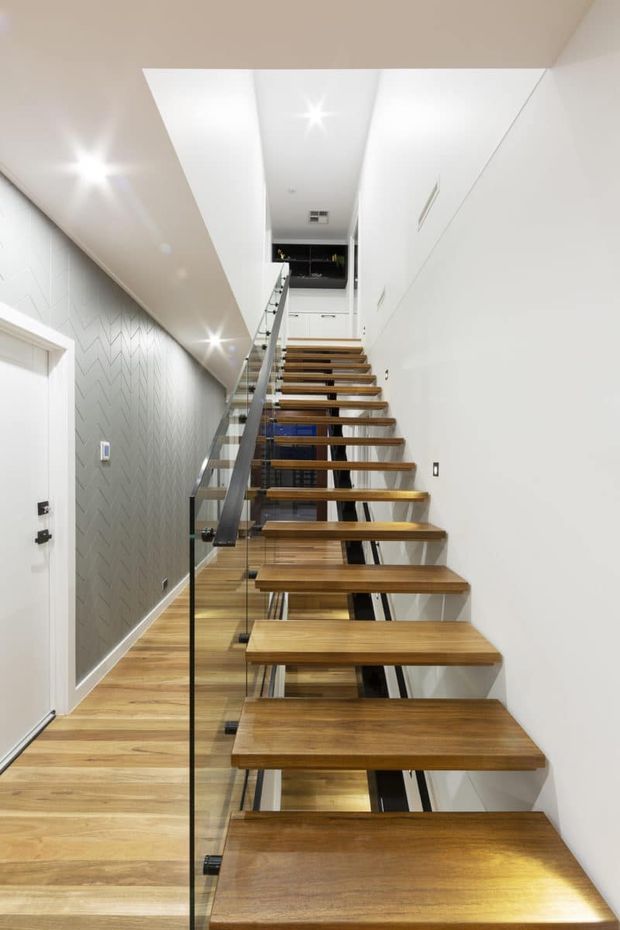 A bright, modern stairway viewed from below, featuring floating timber treads, a clear glass balustrade with dark metal handrail, white walls and ceiling, recessed lighting, light timber flooring, and subtle geometric wall detailing. Built-in white cabinetry and a landing are visible at the upper level.