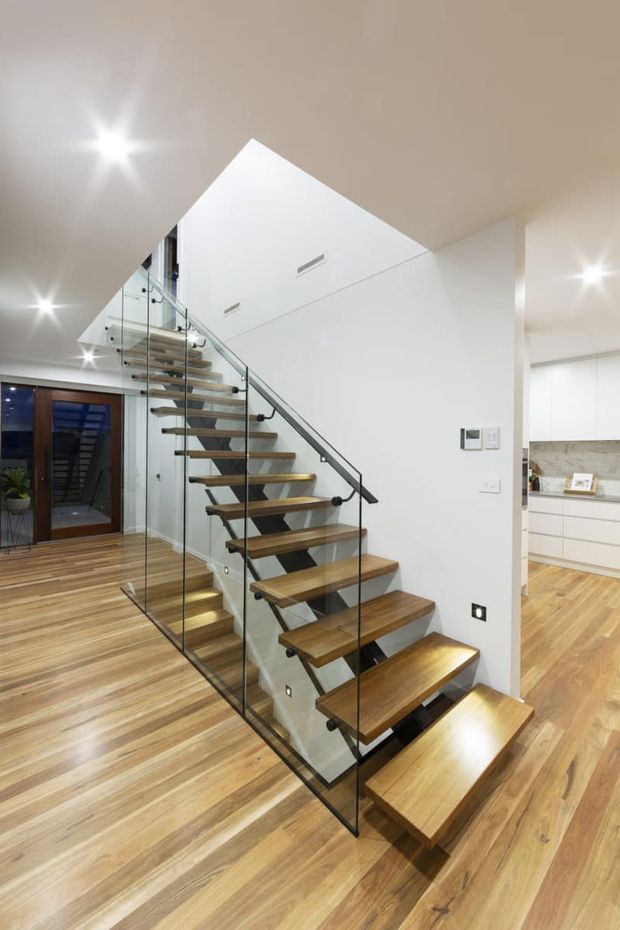 A bright contemporary interior focused on a floating timber staircase with open risers, glass balustrades, and dark metal handrails. The staircase rises through a tall white-walled void, with warm timber flooring continuing into the adjacent kitchen area. Recessed ceiling lights and minimalist white cabinetry reinforce the clean modern design.