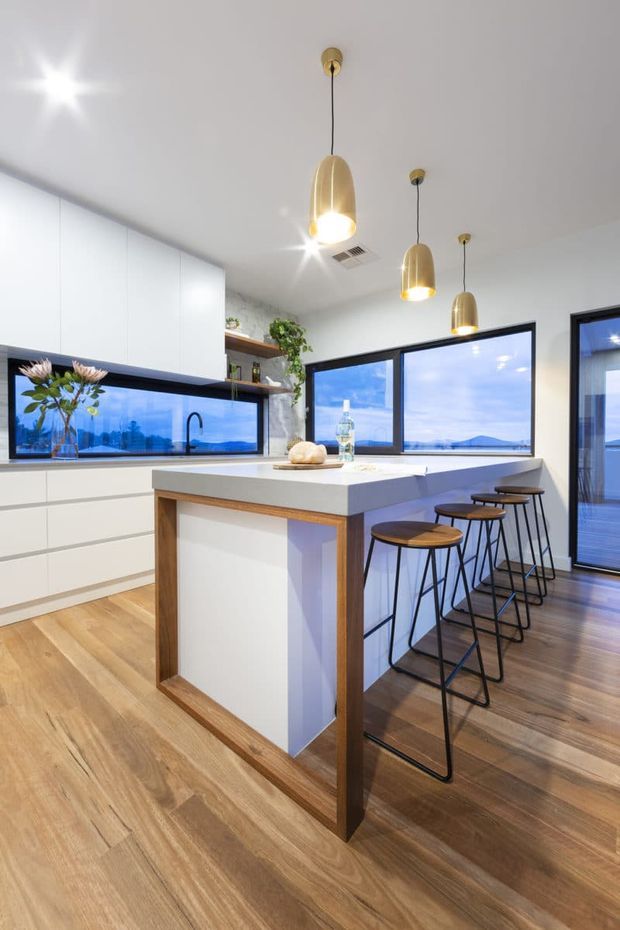 Bright contemporary kitchen with white floor-to-ceiling cabinetry, a large white island with timber edging, warm timber flooring, black-framed windows, and three gold pendant lights. Bar stools line the island, while expansive glazing provides a view of blue water and distant landscape.