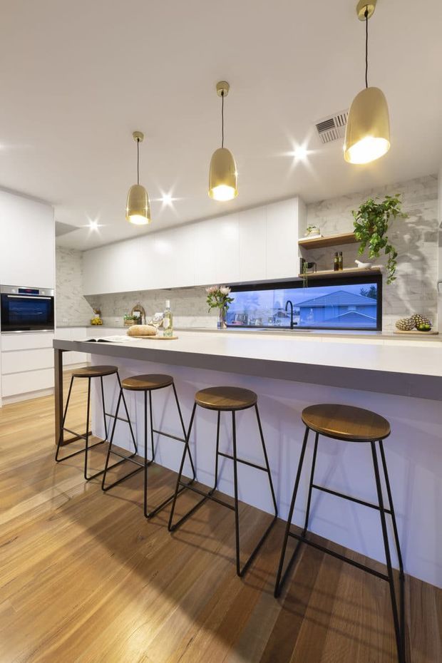 A bright contemporary kitchen with a long white island and integrated breakfast bar seating for four. The kitchen features white full-height cabinetry, a pale marble-look splashback, open timber shelving, warm timber flooring, black-framed glazing, and three gold pendant lights. Minimal styling and clean lines create a spacious, polished interior.