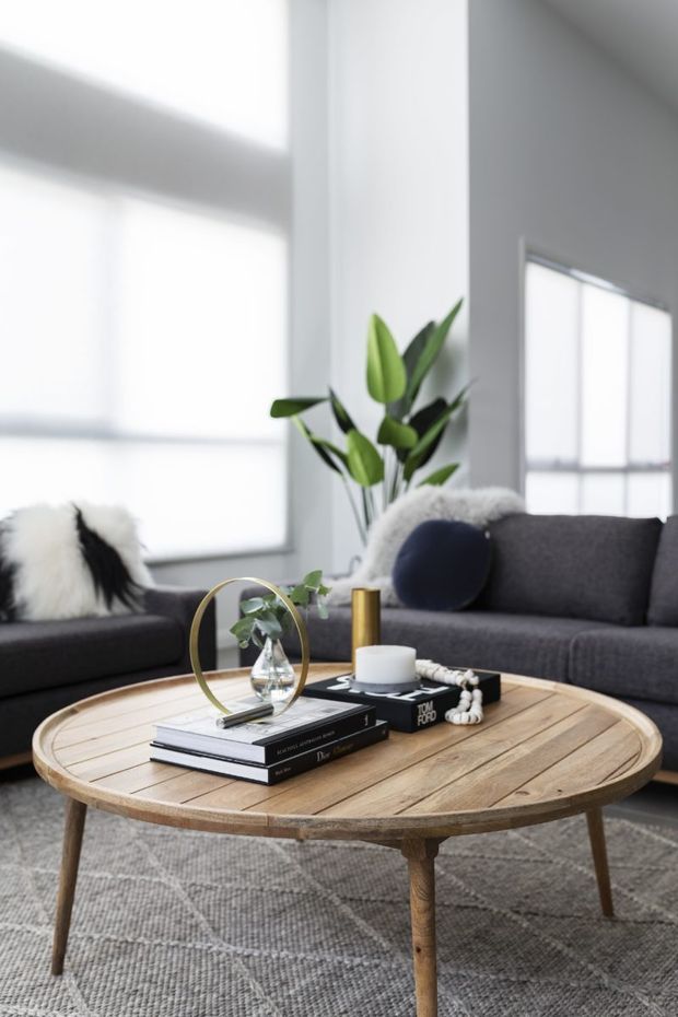 A bright, contemporary living room with a dark grey sectional sofa, textured cushions, a round light timber coffee table, and a neutral geometric rug. Large windows with white roller blinds provide abundant natural light, while a tall indoor plant and carefully arranged books, candle, vase, and decorative accessories add warmth and visual interest.