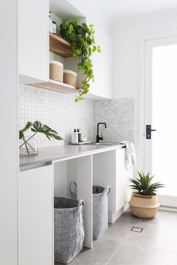 Bright, compact contemporary laundry with white cabinetry, open timber shelving, woven storage baskets, a grey benchtop, black tapware and a geometric white patterned splashback. A large glazed door brings in natural light, while indoor plants add greenery. Grey floor tiles and integrated open storage complete the clean, practical space.