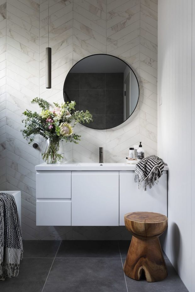 A refined contemporary bathroom featuring a floating white vanity with integrated basin, matte black tapware, a large round black-framed mirror, white marble-look chevron wall tiles, and dark charcoal floor tiles. A pendant light, toiletries, floral arrangement, patterned throw, and sculptural timber stool add decorative detail.