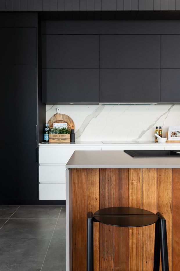 A dark, contemporary kitchen featuring matte charcoal full-height cabinetry, white lower drawers, a white marble-look splashback with grey veining, and a grey benchtop. The island has a warm vertical timber-panelled front with a black stool, creating contrast against the dark cabinetry and large-format dark floor tiles.