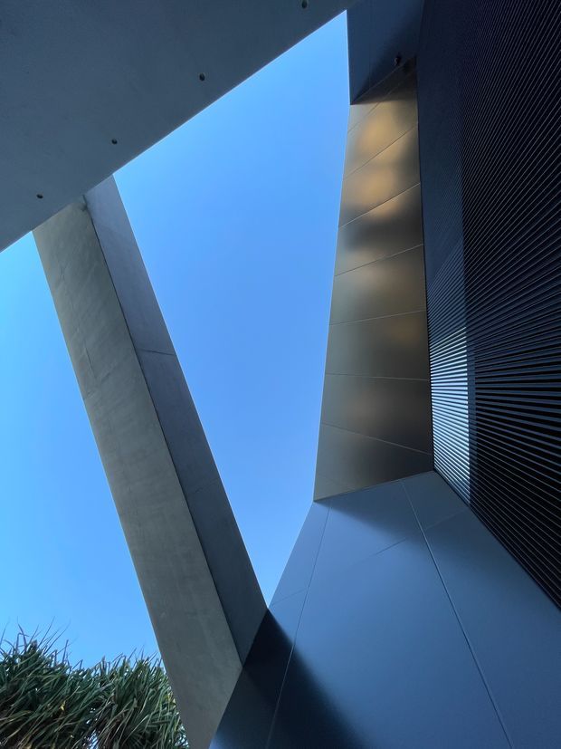 Upward-looking architectural exterior view emphasizing a dramatic angular composition of dark metal cladding, fine vertical louvres, smooth concrete structural elements and a narrow wedge of blue sky. The image highlights geometric lines, a strong cantilevered form and contemporary facade detailing, with minimal landscaping visible at the lower edge.