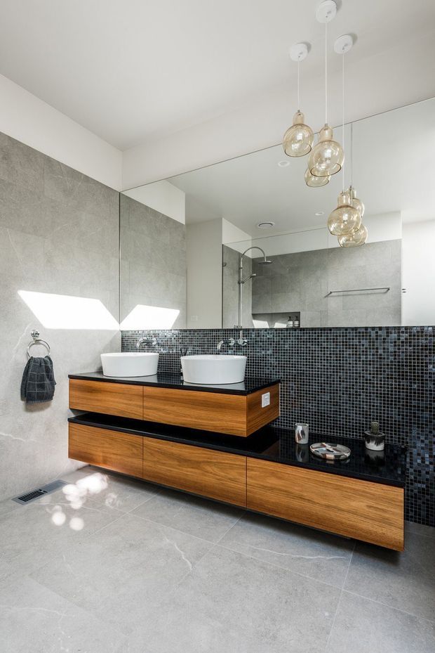 A contemporary bathroom featuring a floating double-vanity with warm timber cabinetry, a polished dark stone benchtop, twin white vessel basins, and a black mosaic tile backsplash. Large-format grey wall and floor tiles, a full-width mirror, and clustered glass pendant lights complete the clean, refined space.