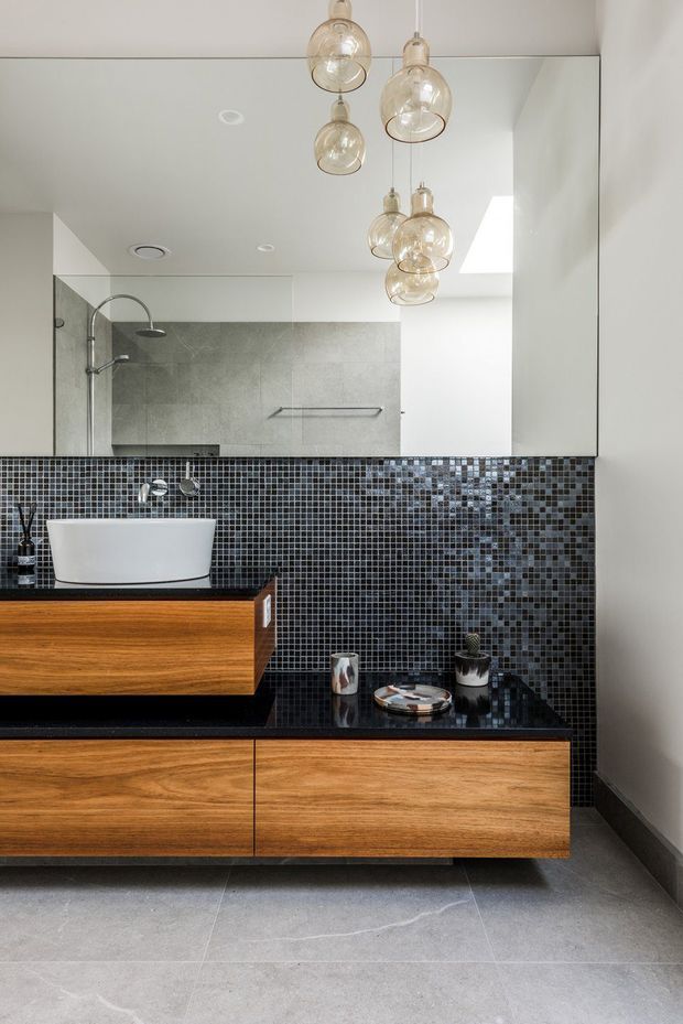 Contemporary bathroom vanity with a freestanding white vessel basin, warm timber drawer cabinetry, black stone benchtops, dark glossy mosaic-tile splashback, and a large mirrored wall. Clear glass shower glazing and a rainfall shower are visible to the left, while multiple suspended glass pendant lights add a decorative focal point.