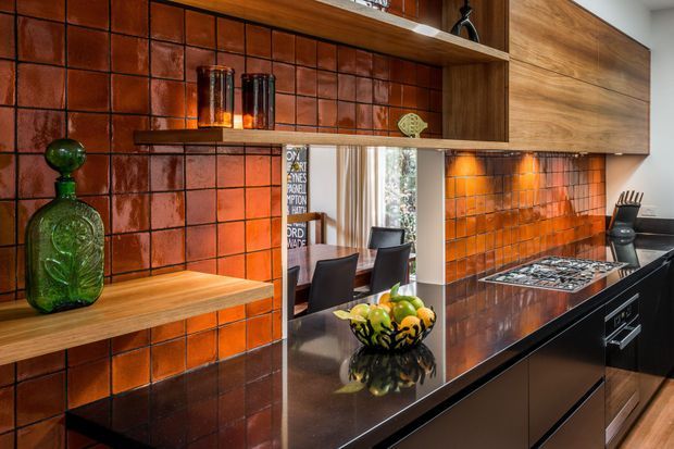 A richly detailed contemporary kitchen featuring a glossy dark stone island benchtop, dark cabinetry, warm timber open shelving and wall cabinetry, and a dramatic burnt-orange glazed tile splashback. A large pass-through opening connects visually to a dining area, while integrated lighting highlights the shelving and benchtop. Decorative glassware and a fruit bowl add colour and texture.
