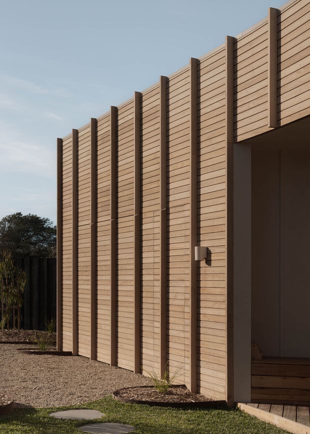 A close exterior view of a contemporary building clad in warm horizontal timber boards, articulated by regularly spaced dark vertical battens. A recessed entry or covered opening is visible at right, opening onto timber steps. The building sits beside a gravel landscape with sparse planting and a stepping-stone path under a clear sky.