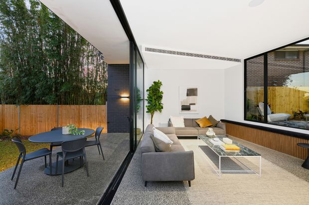 A bright contemporary open-plan living room connected to an outdoor dining terrace through full-height black-framed glazing. The interior features a grey sectional sofa, glass coffee table, pale rug, white walls, timber wall panelling, black brick feature walls, and a large interior window reflecting the adjacent outdoor area. The terrace has a round dining table and chairs, with timber fencing and dense bamboo screening beyond.