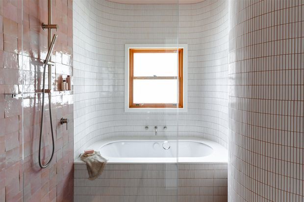 A compact bathroom with a freestanding white bathtub set into a curved alcove, surrounded by small vertically aligned white tiles. The left wall features glossy blush-pink tiles with exposed shower plumbing and a handheld shower, while the window has a warm timber frame. The space has a clean, softly curved, contemporary appearance with natural light.