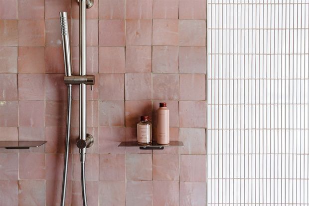 Close-up of a contemporary shower area with dusty pink, irregularly arranged wall tiles, brushed metal shower fittings, slim dark floating shelves holding toiletries, and a white grid-patterned tiled or glazed wall on the right.