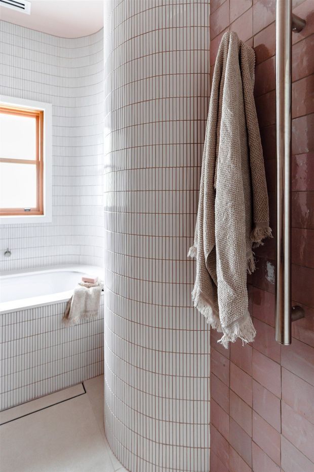 Bright contemporary bathroom with a curved cylindrical wall clad in narrow white tiles, a white bathtub beneath a timber-framed window, and a contrasting dusty-pink tiled wall with a brushed metal towel rail. A textured neutral towel adds warmth and softness.