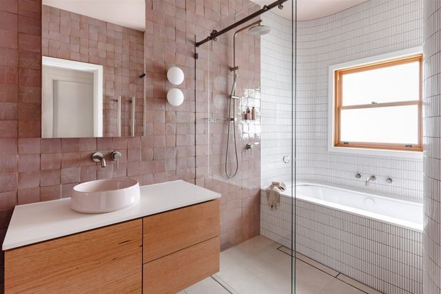 Bright contemporary bathroom featuring a wall-mounted timber vanity with a white vessel basin, white stone benchtop, large-format dusty pink wall tiles, fine white grid tiles around the bathtub, a glass-enclosed walk-in shower, and a timber-framed window. The space has clean lines and warm natural finishes.