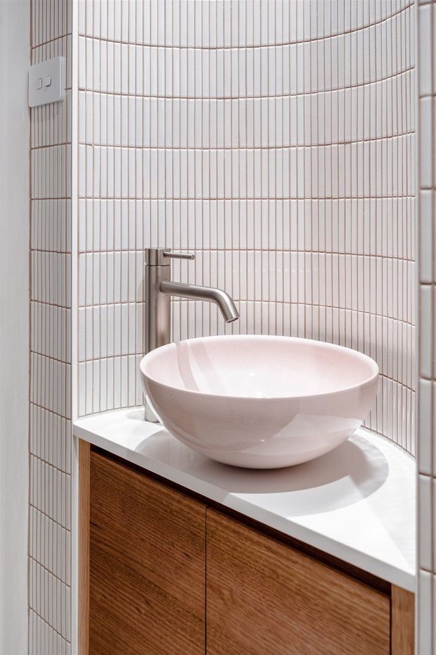 Close-up of a compact contemporary bathroom vanity with a white vessel basin, brushed metal wall-mounted tap, white countertop, and warm timber cabinetry. The walls are clad in narrow vertically ridged white tiles with light grout, wrapping around a curved corner.