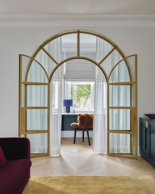 Refined interior view centered on a dramatic arched, glazed timber-and-brass-toned partition with paired glass doors. The partition opens to a bright window-seat workspace or reading nook, with sheer curtains, a compact desk, a rust-brown chair, and a blue table lamp. Pale herringbone timber flooring, white walls with traditional cornice moulding, a deep burgundy sofa edge, a dark teal built-in cabinet, and a muted gold rug create an elegant, contemporary-classic atmosphere.