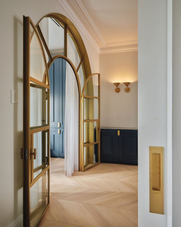 Refined residential entry hall featuring tall arched glazed timber-framed double doors with brass hardware, opening toward a blue paneled door and dark navy built-in cabinetry. The space has warm herringbone timber flooring, pale walls, ornate ceiling cornices, sheer curtains, and paired brass wall sconces, creating a classic yet updated interior.
