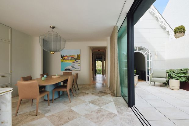 A bright open-plan dining area with a round timber dining table and upholstered chairs, connected to a long hallway and an outdoor tiled courtyard through expansive black-framed sliding glass doors. The interior features large-format patterned stone or marble-look floor tiles, pale walls, a sculptural pendant light, and built-in cabinetry. The courtyard includes a white decorative facade with an arched doorway, greenery, and outdoor seating.