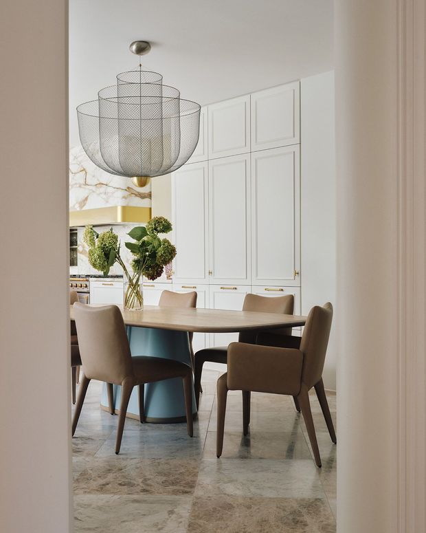 Elegant dining area opening into a refined kitchen, viewed through a doorway. The space features a round or oval timber dining table with upholstered dining chairs, a sculptural oversized wire pendant light, pale blue-green table base, white framed kitchen cabinetry with brass hardware, a veined marble backsplash, and polished stone flooring. A floral arrangement forms the centrepiece, while the overall palette is soft, warm, and sophisticated.