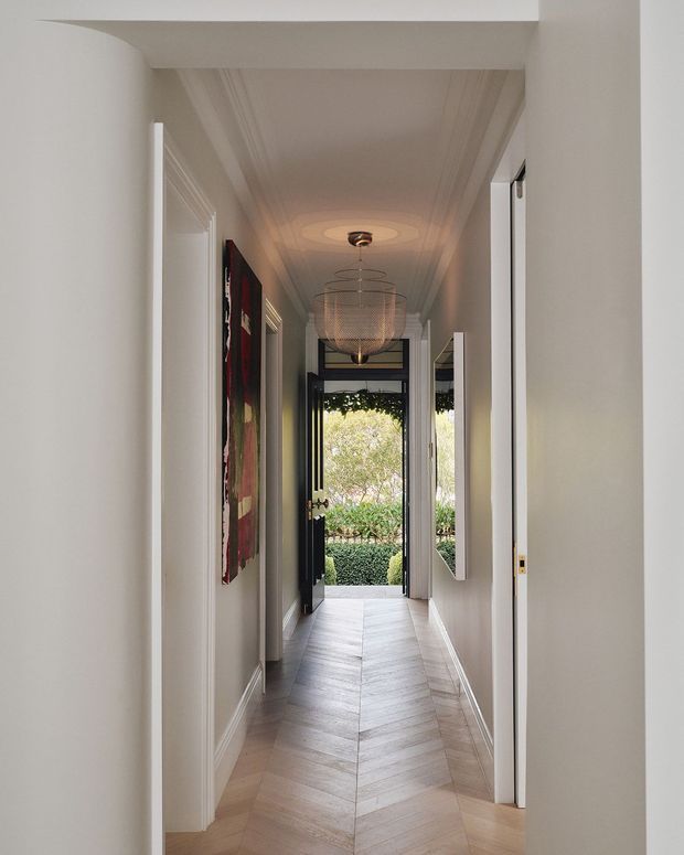 A narrow, elegant residential hallway leading to an open exterior door and garden. The corridor features white walls with decorative trim and cornices, a paneled ceiling, light timber herringbone flooring, white door frames, a tall wall mirror, artwork, and a suspended wire pendant light. Natural light enters from the garden, creating a refined and inviting transitional space.