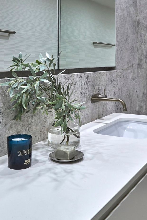 Close-up view of a refined contemporary bathroom vanity with a white solid-surface benchtop, integrated basin, brushed metal wall-mounted tapware, grey stone-look wall finish, and large mirrored cabinetry. Decorative greenery and candles add a soft natural accent.
