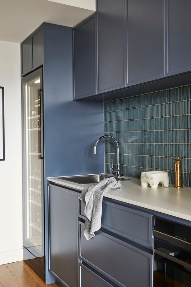 A compact, refined kitchen or butler's pantry zone with full-height navy-blue cabinetry, a pale benchtop, stainless-steel undermount sink and curved chrome tap. The backsplash features vertically arranged teal glazed tiles with contrasting grout. A glass-fronted drinks or storage cabinet is visible at left, while integrated dark appliances occupy the lower right. Timber flooring and minimal decorative accessories complete the space.