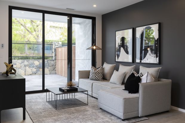 A refined contemporary living room with a large light-grey sectional sofa, black metal-framed glass coffee table, floor-to-ceiling black-framed sliding doors, and a dark charcoal feature wall. Abstract framed artwork, neutral cushions, a sculptural floor lamp, and a textured rug create a monochromatic, polished interior. The glazing opens to a paved outdoor courtyard with a stone retaining wall and timber gate.