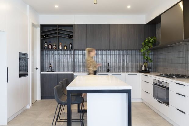 Contemporary kitchen with a large light-coloured island and dark bar seating, extensive dark timber-toned cabinetry, a full-height ribbed grey splashback, integrated appliances, open bottle shelving, and a gas cooktop. The bright, streamlined space has a clean monochromatic palette with warm wood accents.