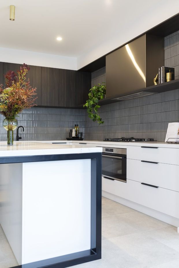 A contemporary kitchen with white flat-panel cabinetry, dark timber overhead joinery, a dark vertically ribbed tile splashback, pale stone benchtops, and a large island with a dark frame and illuminated white panel. Integrated appliances, gas cooktop, open shelving, indoor plants, and warm decorative lighting create a refined high-contrast interior.