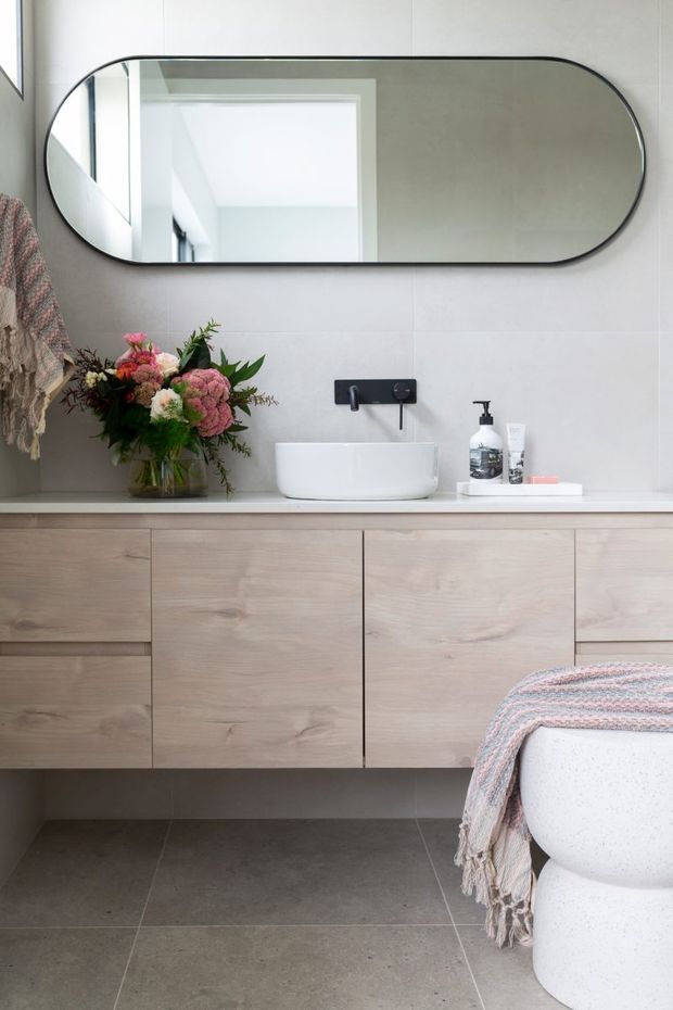 A refined contemporary bathroom featuring a wall-mounted light timber vanity with integrated drawers, a white vessel basin, matte black wall-mounted tapware, and a large rounded rectangular mirror. Pale tiled walls and large grey floor tiles create a calm neutral backdrop, with a freestanding white bath partially visible at right and soft floral and textile accents.