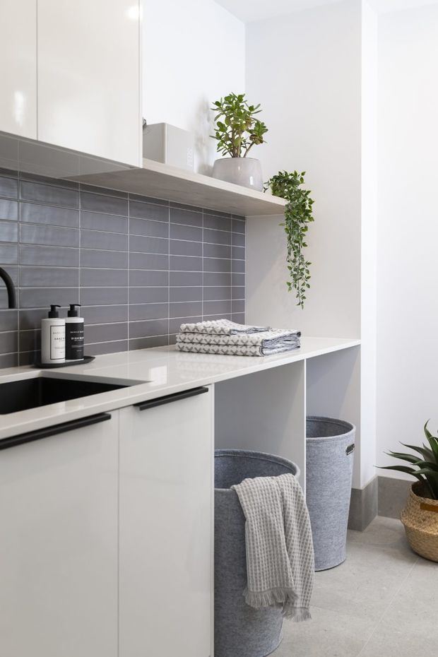 Bright, compact laundry room with white built-in cabinetry, a pale benchtop, dark grey rectangular tiled splashback, open shelving, folded towels, laundry hampers, and potted greenery. The space has a clean contemporary look with light grey flooring and black hardware.