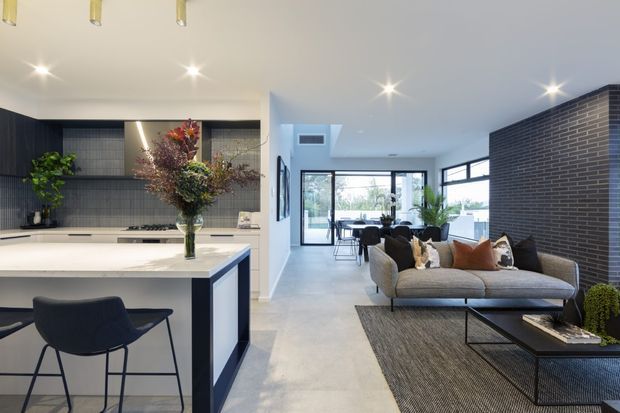 Open-plan contemporary interior combining a kitchen, dining area and living room. The kitchen features a large light stone island with dark cabinetry and a tiled splashback, while the living area has a grey sectional sofa, black coffee tables and a dark brick feature wall. Full-height glazing at the rear brings in abundant daylight and connects the space to an exterior view.